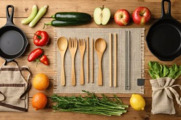 An assortment of eco-friendly kitchen utensils including bamboo spoons, stainless steel straws, silicone baking mats, and cast iron skillets displayed on a wooden countertop with fresh produce.