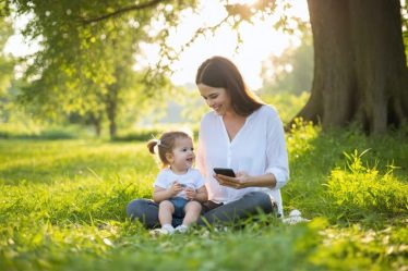 A parent and child sitting cross-legged in a grassy field, smiling at each other, with a phone placed aside, depicting the theme of connection and mindfulness.