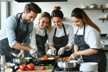 A diverse team of corporate professionals enjoying a hands-on cooking class, collaborating and smiling while preparing dishes in a professional kitchen setting.