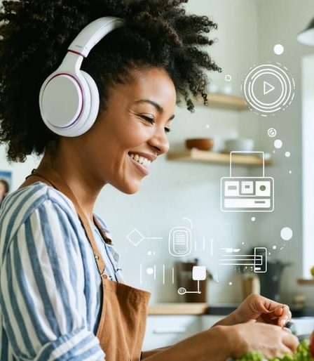 A parent smiling and listening to a podcast with headphones while multitasking in the kitchen, depicting the integration of podcast learning into daily family life.