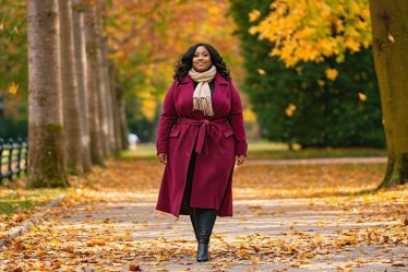 Plus-size model in a jewel-toned wool coat confidently walking in a picturesque fall park, surrounded by colorful autumn leaves, highlighting sophisticated layered fashion.