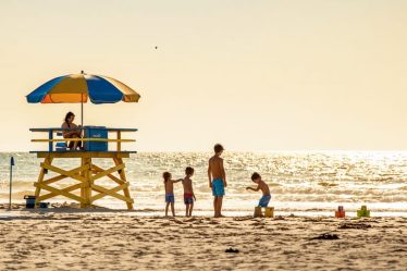 Calm beach setting with a lifeguard tower, where a family enjoys building sandcastles and playing in shallow, gentle waters while parents relax under an umbrella.