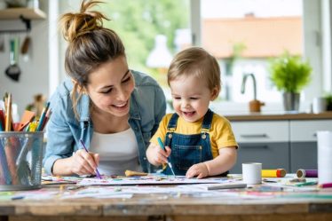 Mother with a messy bun engaging in a craft activity with her toddler in a lively, disorderly home setting, illustrating the everyday chaos and joy of parenting.