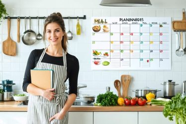 A busy mom standing in a well-organized kitchen, holding a meal planning notebook with various fresh ingredients on the counter and a calendar of themed dinner nights on the wall.