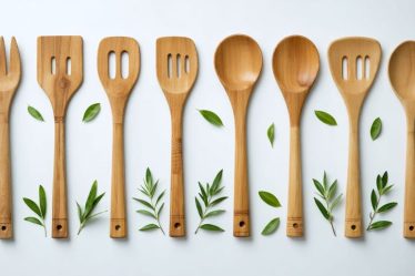 Assorted eco-friendly kitchen utensils including bamboo spatulas, wooden spoons, and reclaimed wood tools arranged against a light neutral background with hints of greenery.