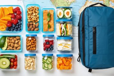 Assorted travel-friendly meal prep items laid out on a table, including containers with mixed nuts, dried fruits, protein bars, wrap sandwiches, and mason jar salads, set against a background featuring a travel map and a mini fridge.