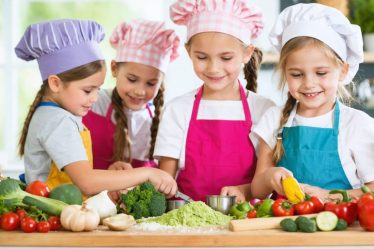 Energetic children wearing colorful aprons and chef hats enthusiastically participating in a hands-on cooking class, learning kitchen skills with fresh ingredients in a lively, bright kitchen setting.