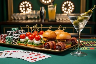 Close-up of a green-felt poker table with cards, stacked chips, a platter of caprese skewers, mini sliders, bacon-wrapped dates, and a martini; a self-serve cocktail station is softly blurred in the background.