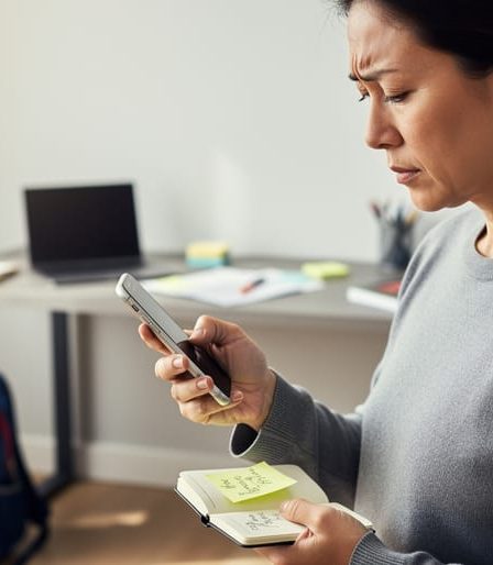 Parent seated at home holding a smartphone and notebook while pausing to regroup, with a blurred laptop and school items in the background.